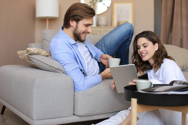 Feliz pareja joven sentada, relajándose en el piso en la sala de estar viendo contenido multimedia en línea, haciendo compras en línea en una tableta.