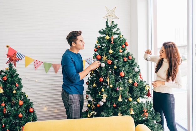 Feliz pareja joven usando suéteres divirtiéndose decorando varios adornos en el árbol de Navidad en la sala de estar en casa Disfrutando y celebrando las vacaciones de Navidad