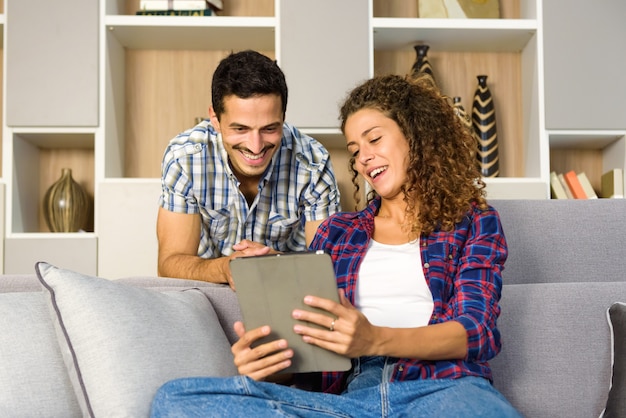 Feliz pareja joven viendo los medios de comunicación en una tableta juntos en la sala de estar en casa sonriendo con placer y disfrute