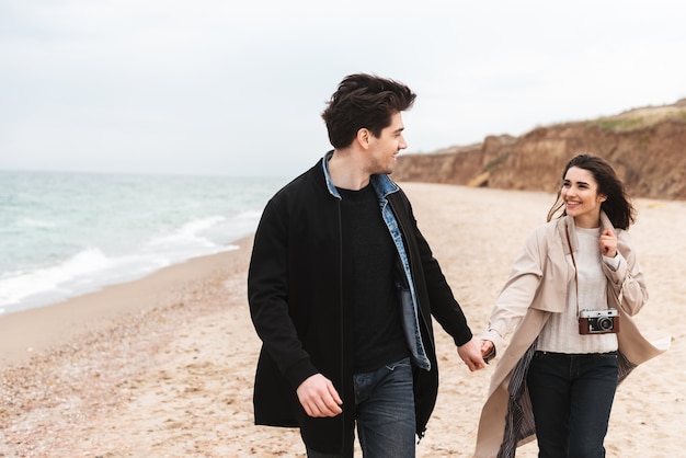 Feliz pareja joven vistiendo abrigos de otoño pasar tiempo juntos en la playa, caminando