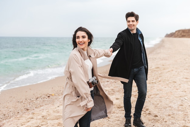 Feliz pareja joven vistiendo abrigos de otoño pasar tiempo juntos en la playa, caminando