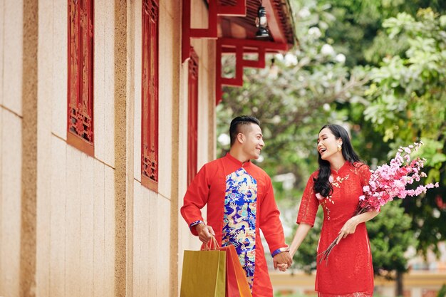 Feliz pareja de jóvenes asiáticos cogidos de la mano al caminar al aire libre en ropa tradicional con ramas florecientes y bolsas de la compra.