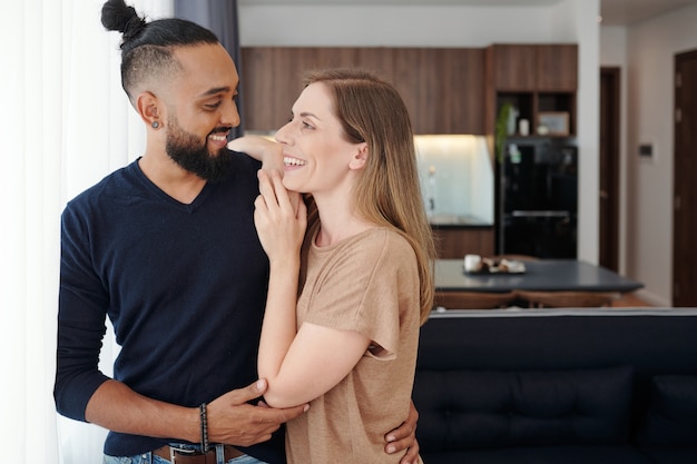 Foto feliz pareja de jóvenes de raza mixta en el amor abrazándose y mirándose cuando está de pie en el apartamento estudio