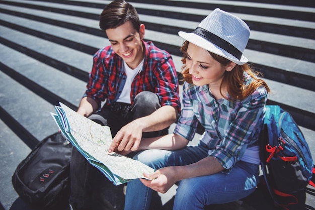 Feliz pareja de jóvenes turistas sentados juntos en las escaleras y examinando el mapa.