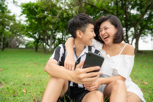 Feliz pareja de lesbianas asiáticas riendo sentado al aire libre en el parque mientras usa el teléfono móvil