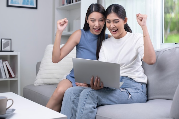Feliz pareja de lesbianas asiáticas usando una laptop en la sala de estar felizmente levantando sus manos celebrando el éxito ganador o las buenas noticias