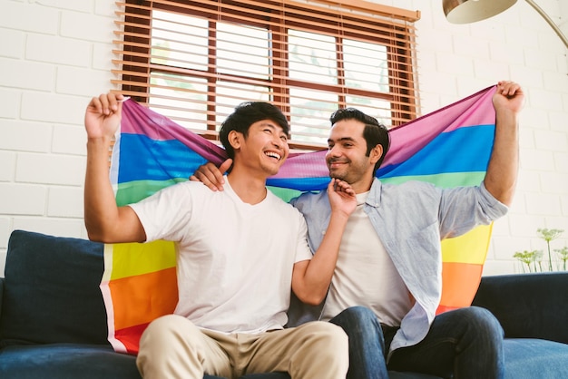 Feliz pareja LGBT caucásica y asiática sentada en el sofá sosteniendo y ondeando la bandera del orgullo LGBT del arco iris juntos en la sala de estar en casa Diversidad de las relaciones LGBT Un concepto de pareja gay