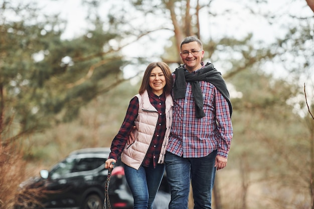 Feliz pareja madura da un paseo por el bosque de otoño o primavera cerca de un coche moderno