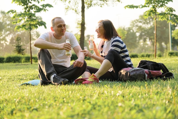 Feliz pareja madura sentada en el parque en la estera de fitness, descansando