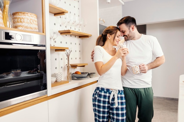 Una feliz pareja matutina con café abrazándose en la cocina de su acogedor apartamento