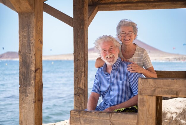 Feliz pareja mayor caucásica sentada a la sombra de la glorieta frente al mar mirando a la cámara riéndose de los ancianos jubilados disfrutando de las vacaciones en el mar en un día ventoso y soleado