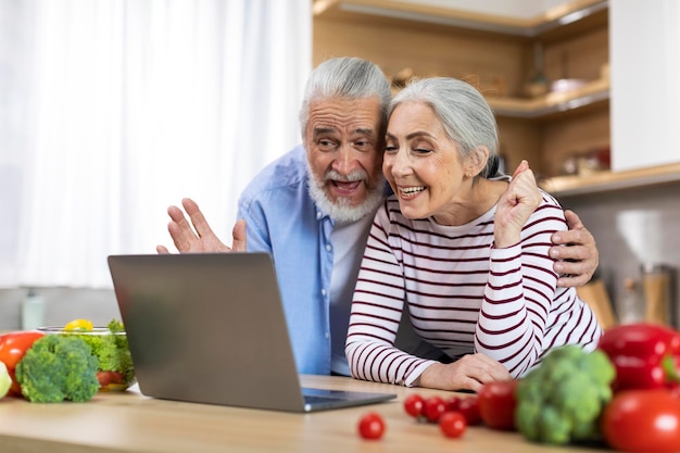 Feliz pareja mayor haciendo videollamadas con una laptop mientras se relaja en la cocina