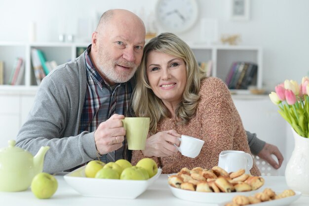 Feliz pareja mayor sentada en la mesa de la cocina y bebiendo té con galletas