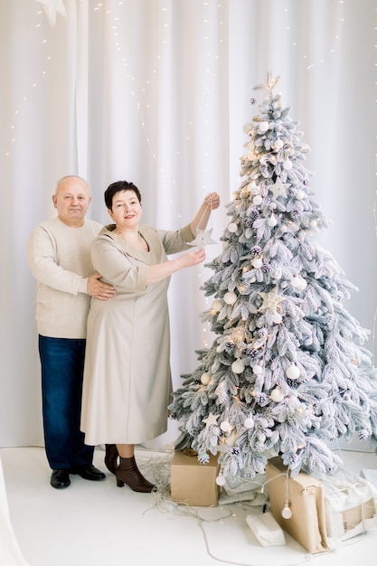 Feliz pareja de mediana edad preparándose para las vacaciones de invierno decorando el hermoso árbol de Navidad en el acogedor estudio de luz