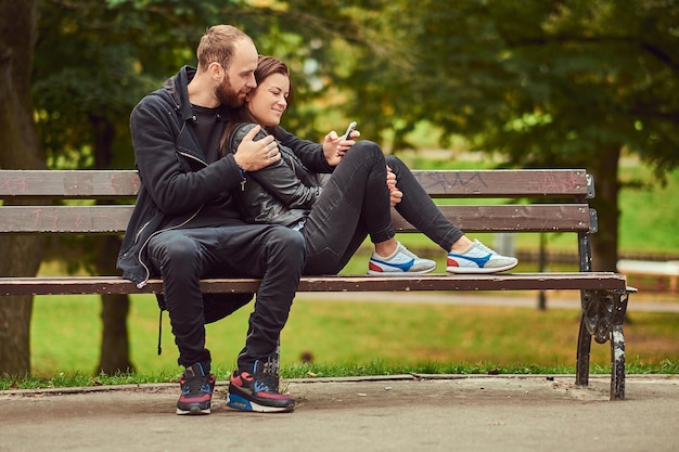Feliz pareja moderna, buscando algo divertido en Internet mientras se abraza en un banco en el parque. Disfrutando de su amor y la naturaleza.