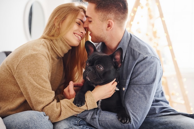 Feliz pareja moderna joven sentada en el sofá en casa con árbol de Navidad con su lindo perro.