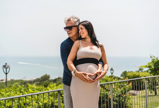 Feliz pareja con una mujer embarazada de pie frente a un hermoso paisaje marino durante las vacaciones de verano Hermosa esposa y un hombre de mediana edad que se ríen juntos esperando un bebé Concepto de embarazo
