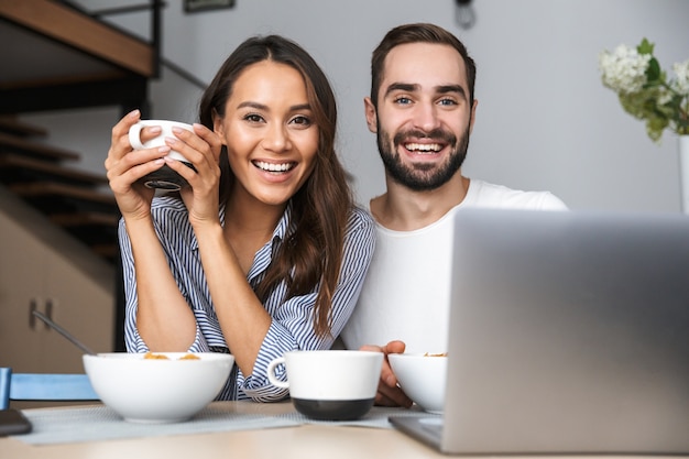 Feliz pareja multiétnica desayunando en la cocina, mirando la computadora portátil