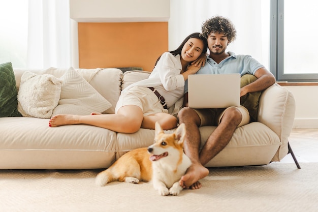 Una feliz pareja multiétnica relajándose en el sofá con el perro y usando una computadora portátil