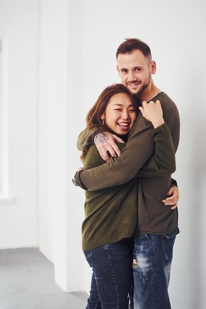 Feliz pareja multiétnica con ropa informal abrazándose unos a otros en el interior del estudio. Chico caucásico con novia asiática.