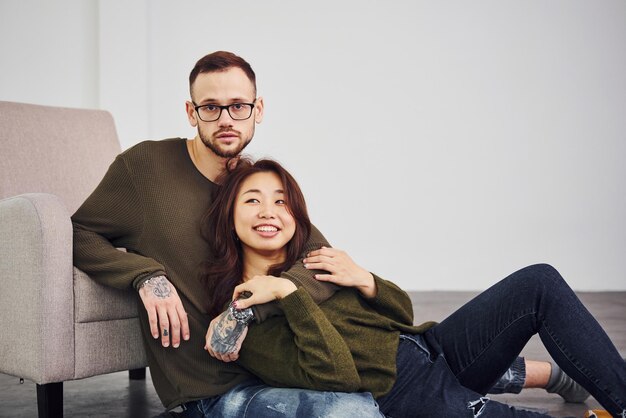 Feliz pareja multiétnica con ropa informal sentada en el interior del estudio. Chico caucásico con novia asiática.