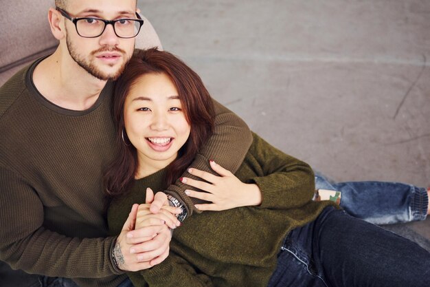 Feliz pareja multiétnica con ropa informal sentada en el interior del estudio. Chico caucásico con novia asiática.