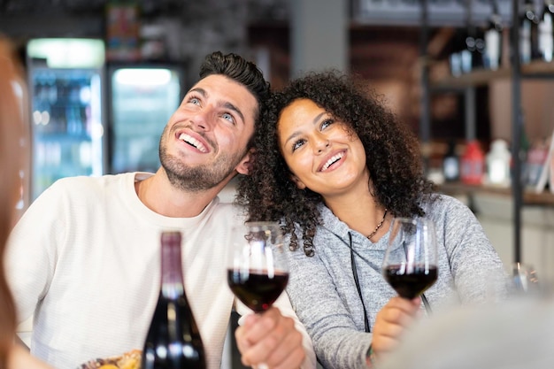 Feliz pareja multirracial de amantes bebiendo vino tinto en el restaurante de moda Situación divertida con hombre y mujer mirando hacia arriba Concepto de relación con novio y novia en la primera cita