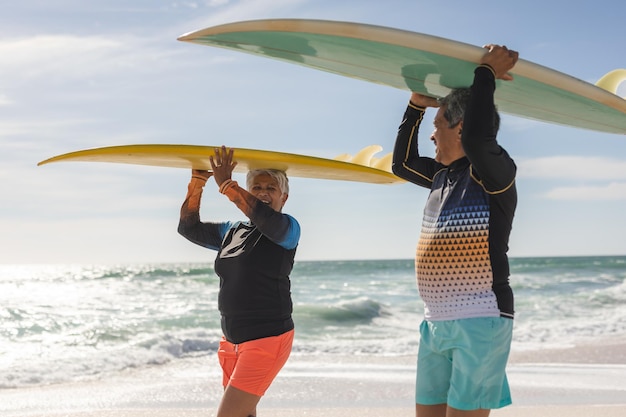 Feliz pareja multirracial de ancianos mirándose mientras llevaban tablas de surf en la cabeza en la playa