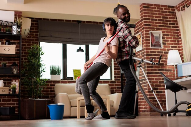 Feliz pareja multirracial bailando cantando aspirando y trapeando el piso de la sala de estar en una casa moderna. Hombre y mujer emocionados divirtiéndose mientras terminan la limpieza de primavera en casa.