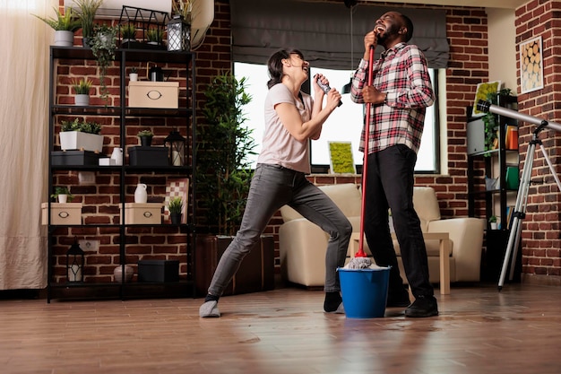 Feliz pareja multirracial cantando con emoción, disfrutando juntos de las tareas domésticas. Mujer cantando con control remoto mientras el esposo usa un trapeador como micrófono, haciendo limpieza de primavera con buenas vibraciones.