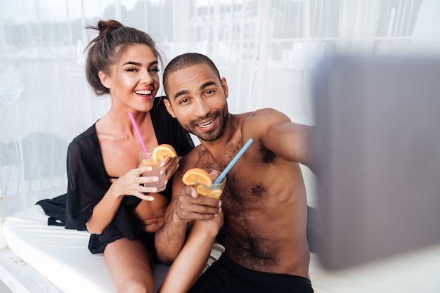 Feliz pareja multirracial sonriente haciendo selfie y sosteniendo cócteles en la playa