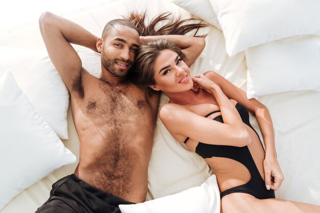 Feliz pareja multirracial tendido en la cama de playa blanca y mirando a la cámara