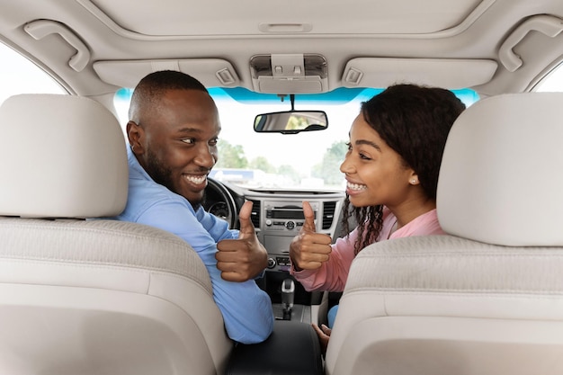 Feliz pareja negra sentada en un auto gesticulando con los pulgares hacia arriba