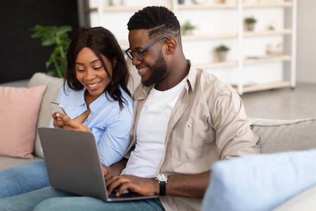 Feliz pareja negra sentada en el sofá usando celular y pc