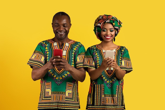 Feliz pareja negra con trajes africanos usando teléfonos inteligentes