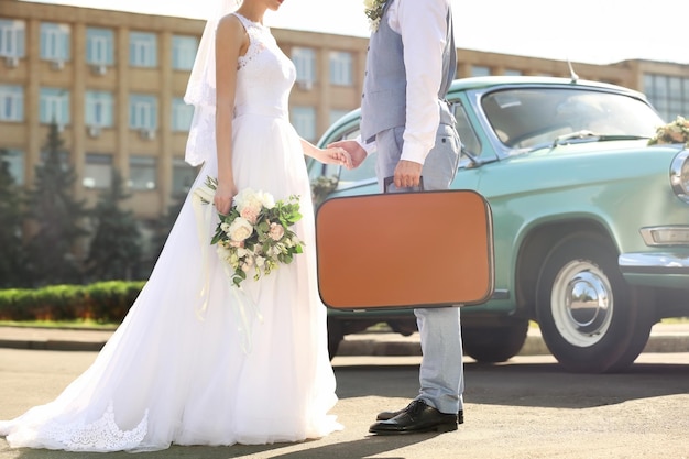 Feliz pareja de novios con maleta y coche al aire libre