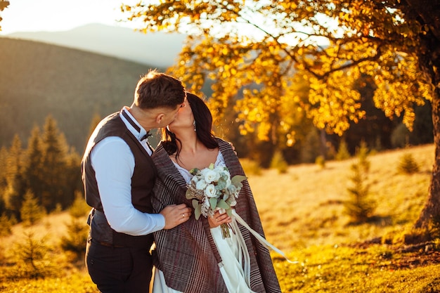 Feliz pareja de novios quedándose y besándose en el hermoso paisaje con montañas durante la puesta de sol