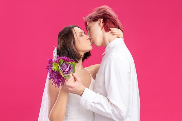 Feliz pareja de novios en vestido de novia con flores feliz enamorado abrazándose y besándose de pie sobre la pared rosa