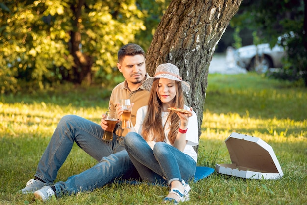 Feliz pareja de picnic en el parque. Comiendo pizza y bebiendo cerveza