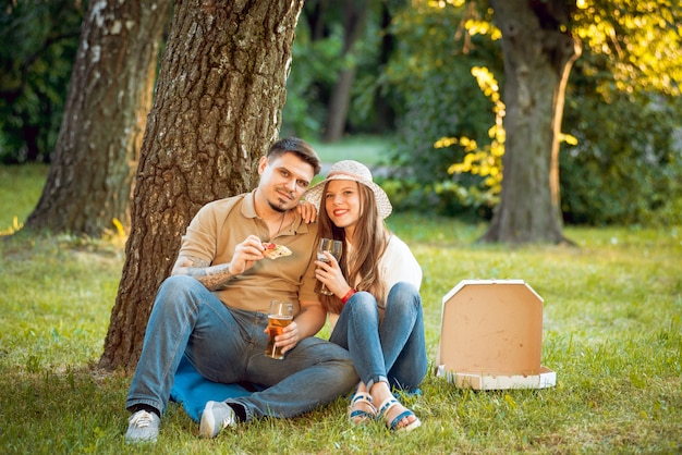 Feliz pareja de picnic en el parque. Comiendo pizza y bebiendo cerveza