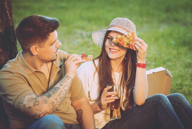 Feliz pareja de picnic en el parque. Comiendo pizza y bebiendo cerveza