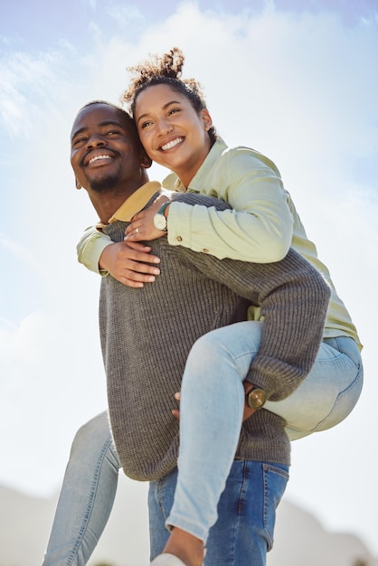 Feliz pareja y piggy back disfrutan de aire fresco en vacaciones al aire libre con fondo de cielo azul Sonrisa hombre negro y mujer feliz siendo juguetones y divirtiéndose juntos en una cita romántica en el parque natural