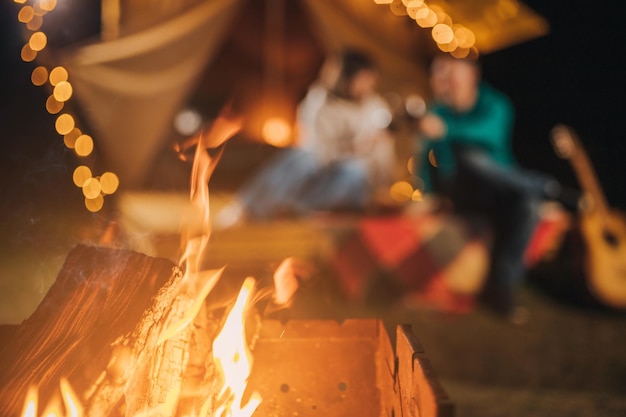 Feliz pareja relajándose en glamping en la noche de otoño bebiendo vino y tocando la guitarra cerca de una acogedora hoguera Tienda de campaña de lujo para recreación al aire libre y recreación Concepto de estilo de vida