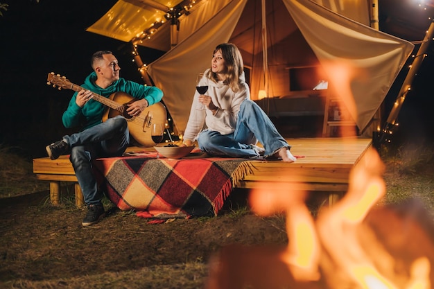 Feliz pareja relajándose en glamping en la noche de otoño bebiendo vino y tocando la guitarra cerca de una acogedora hoguera Tienda de campaña de lujo para recreación al aire libre y recreación Concepto de estilo de vida