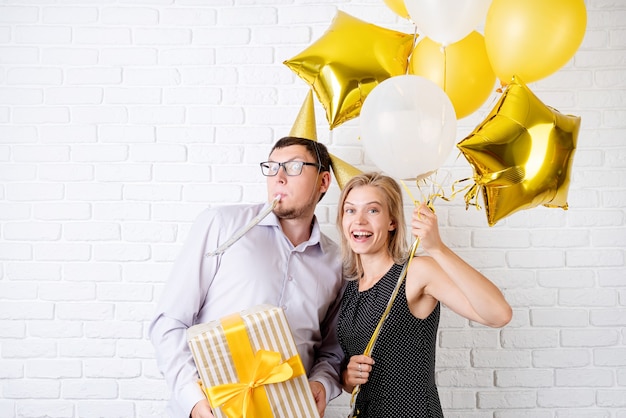 Foto feliz pareja riendo celebrando la fiesta de cumpleaños con globos dorados y caja de regalo sobre pared de ladrillo blanco.