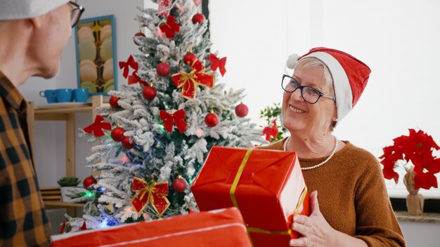 Feliz pareja senior disfrutando de Navidad compartiendo envoltorio regalo presente en cocina decorada de Navidad