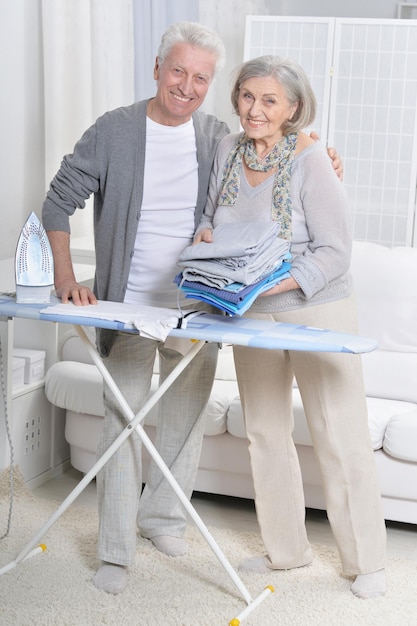 Feliz pareja senior posando y durante el planchado en casa