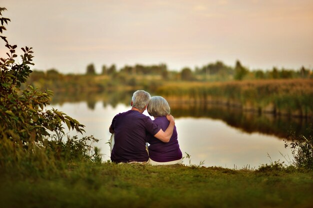 Feliz pareja senior sentada en verano cerca del lago durante la puesta de sol, vista posterior