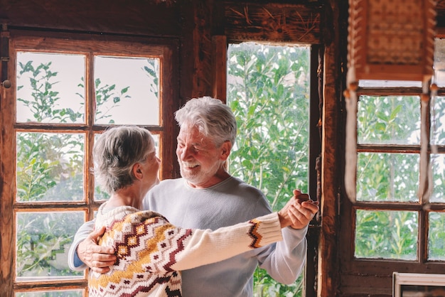 Feliz pareja senior sonríe y baila en casa disfrutando del amor y la relación juntos. El anciano activo y la mujer se divierten en la actividad de ocio interior. Vista del bosque de la naturaleza desde windows