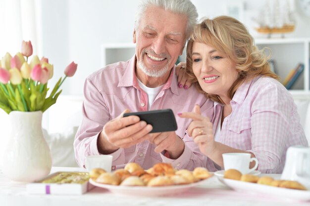 Feliz pareja senior con teléfono inteligente bebiendo té en la cocina